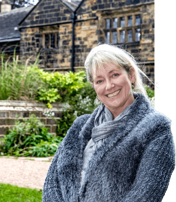 Angela, wearing a fluffy grey cardigan and scarf, stands smiling in front of Elizabethan manor house, Oakwell Hall.