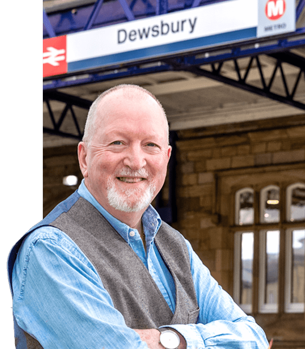 Edward, wearing a blue shirt and grey waistcoat, sits smiling with arms crossed outside Dewsbury train station.