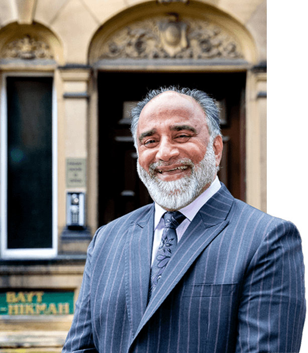 Nadeem, wearing a suit, tie and purple shirt stands smiling, framed by the large doorway of the Al-Hikmah centre in Batley.